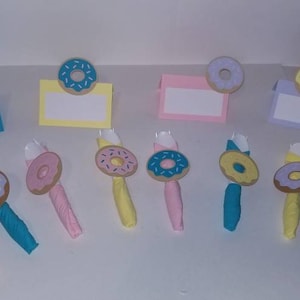 May include: A set of donut-themed place card holders and napkin rings. The place card holders are made of cardstock and feature a donut with sprinkles in pink, blue, and yellow. The napkin rings are made of fabric and feature a donut with sprinkles in pink, blue, and yellow.