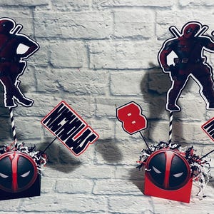 May include: Two red and black Deadpool themed centerpieces with the name Nicholas and the number 8 on them. Each centerpiece has a red and black Deadpool mask on a small box.