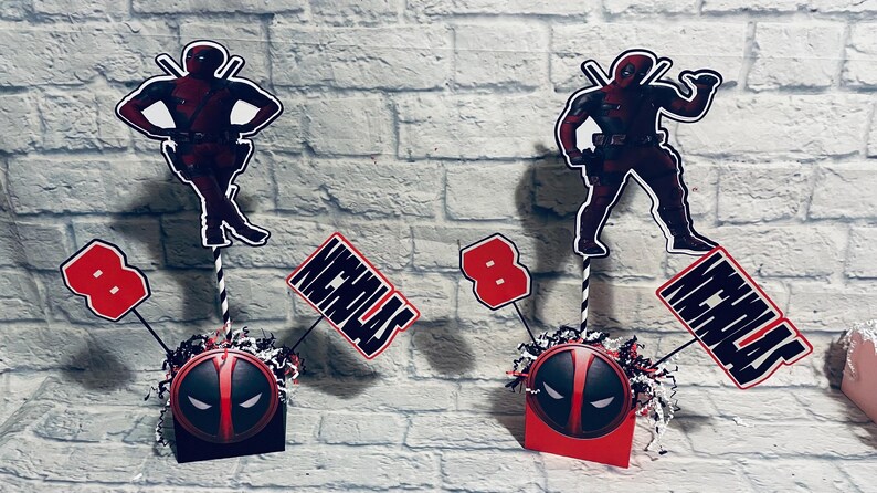 May include: Two red and black Deadpool themed centerpieces with the name Nicholas and the number 8 on them. Each centerpiece has a red and black Deadpool mask on a small box.