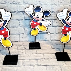 May include: Three Mickey Mouse centerpieces, each featuring a cartoon image of Mickey Mouse in his signature red shorts and yellow shoes, with a black base.