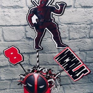 May include: A red and black Deadpool themed centerpiece with a Deadpool figure, a red number 8, and a sign that says "Nicholas".