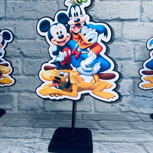 May include: A black wooden stand with a colorful cardboard cutout of Mickey Mouse, Donald Duck, Goofy, and Pluto. The characters are smiling and appear to be hugging.