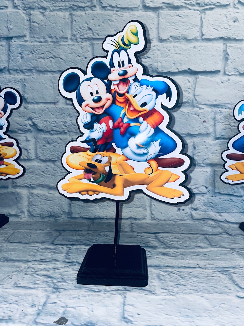 May include: A black wooden stand with a colorful cardboard cutout of Mickey Mouse, Donald Duck, Goofy, and Pluto. The characters are smiling and appear to be hugging.