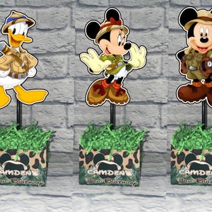 Puede incluir: Cinco centros de mesa con los personajes de Disney Daisy Duck, Donald Duck, Minnie Mouse, Mickey Mouse y Goofy vestidos con ropa de safari. Cada personaje est&aacute; montado en un palo negro en una caja con estampado de leopardo y el nombre CAMDENE.