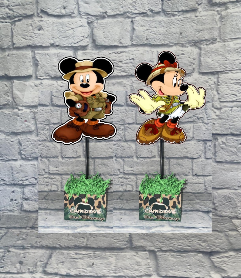 Puede incluir: Dos centros de mesa decorativos con Mickey y Minnie Mouse en traje de safari, montados en un palo negro dentro de cajas con papel verde. Las cajas tienen un estampado de leopardo y el texto CAMDEN'S 3RD BIRTHDAY.