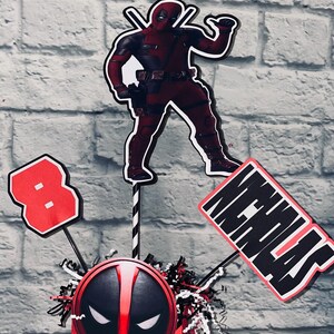 May include: A red and black Deadpool themed centerpiece with a red box featuring a Deadpool mask, a red number 8, a red and black Deadpool character cutout, and a red and black sign that says "Nicholas".