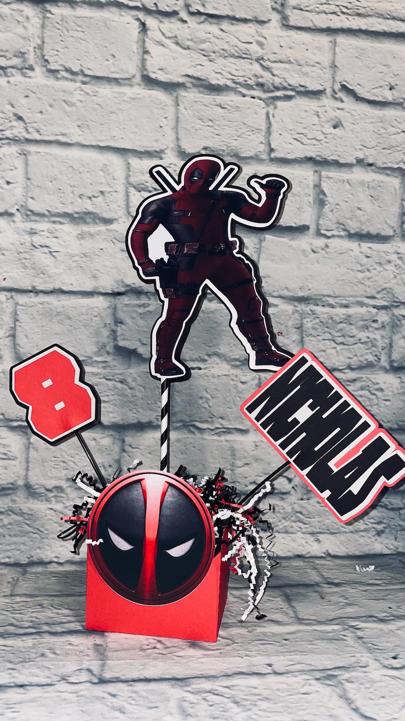 May include: A red and black Deadpool themed centerpiece with a red box featuring a Deadpool mask, a red number 8, a red and black Deadpool character cutout, and a red and black sign that says "Nicholas".