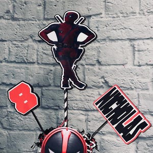 May include: A red and black Deadpool themed centerpiece with a Deadpool character cutout, a red number 8, and a sign that says "Happy Birthday".
