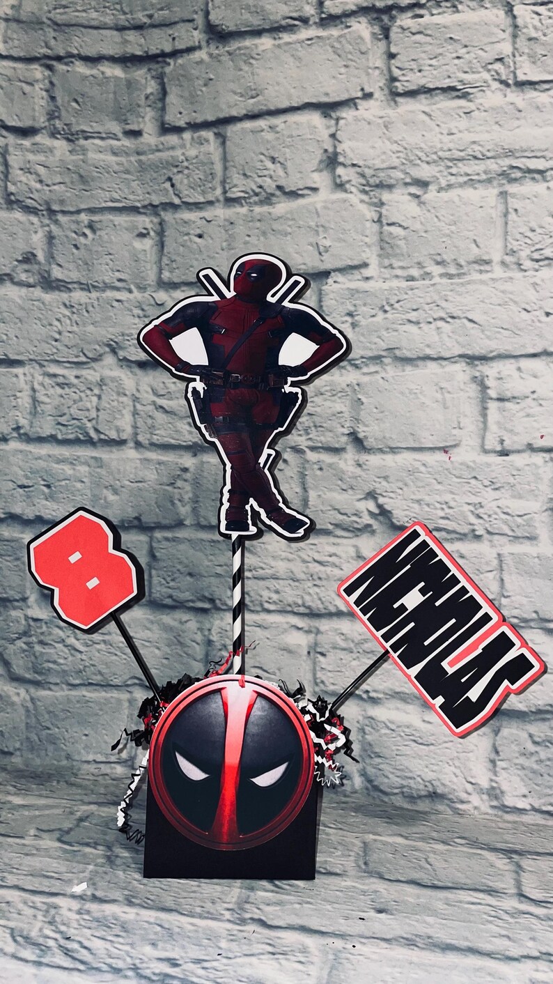 May include: A red and black Deadpool themed centerpiece with a Deadpool character cutout, a red number 8, and a sign that says "Happy Birthday".