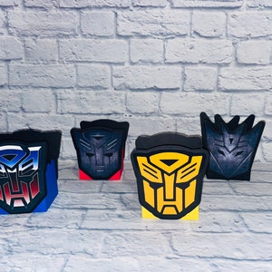 May include: Five cardboard boxes with different Transformers character logos. The boxes are black, blue, red, yellow, and gray. The logos are printed on the front of the boxes.