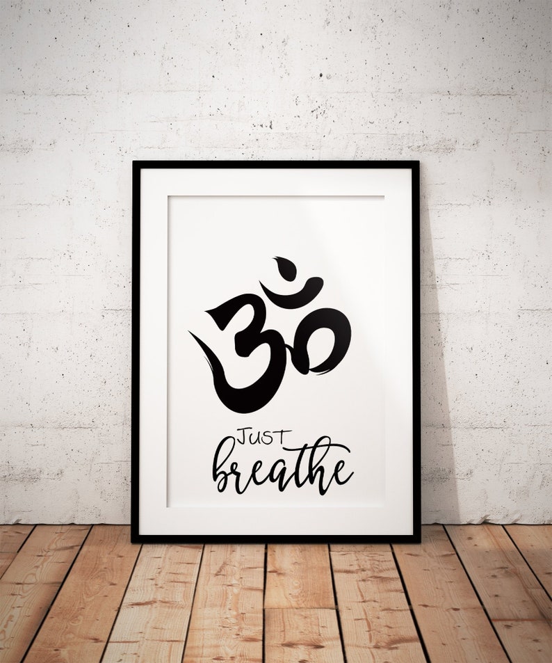 May include: Black framed print featuring a black Om symbol and the words "Just breathe" in a cursive font on a white background. The print is displayed against a white textured wall and a wooden floor.