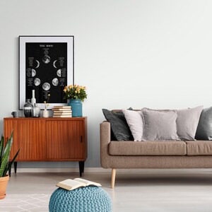 May include: A framed black and white poster titled "THE MOON" displays the phases of the moon against a starry background. A wooden cabinet with vases, books, and a blue vase with flowers sits below the poster. A brown sofa with pillows is to the right.