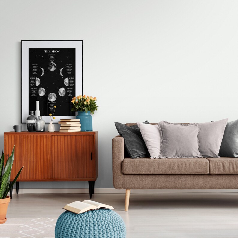May include: A framed black and white poster titled "THE MOON" displays the phases of the moon against a starry background. A wooden cabinet with vases, books, and a blue vase with flowers sits below the poster. A brown sofa with pillows is to the right.