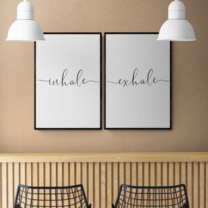 May include: Two black framed prints with white backgrounds and black cursive text. The left print says "inhale" and the right print says "exhale". The prints are hanging on a beige wall above a wooden counter with two black metal chairs.