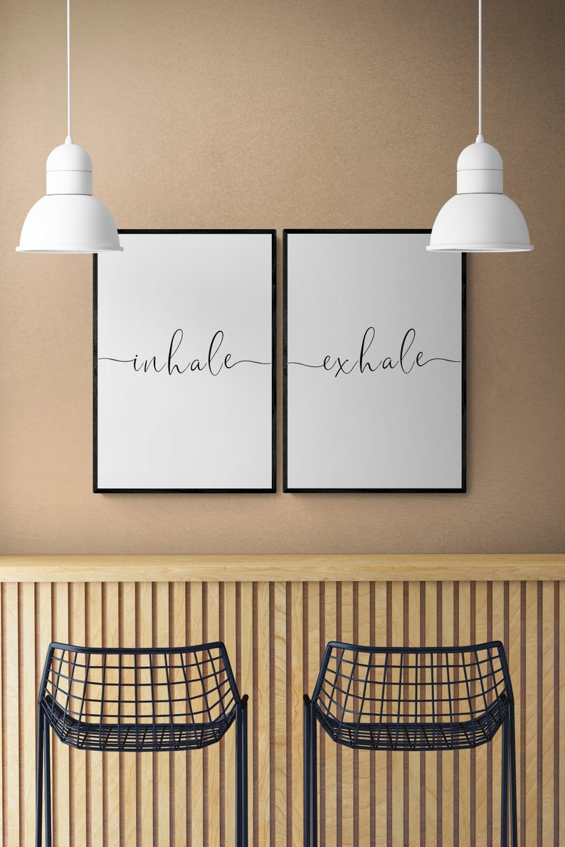 May include: Two black framed prints with white backgrounds and black cursive text. The left print says "inhale" and the right print says "exhale". The prints are hanging on a beige wall above a wooden counter with two black metal chairs.