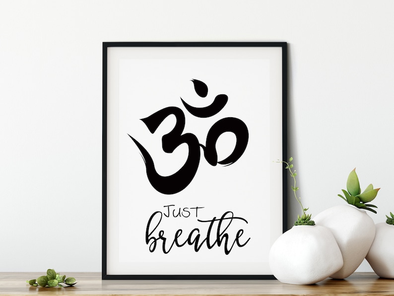 May include: Black framed art print featuring the Om symbol and the words "Just breathe" in a cursive font. The artwork is displayed on a wooden surface, with white vases and greenery in the background.