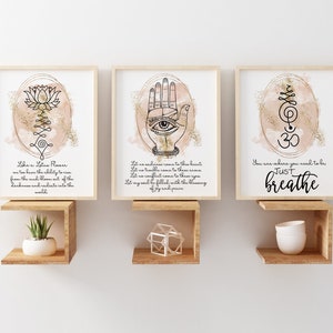 May include: Three framed prints with watercolor backgrounds and gold glitter accents. The first print features a lotus flower with the text "Like a Lotus Flower we too have the ability to rise from the mud, blossom out of the darkness and radiate into the world". The second print features a hand with an eye in the palm and the text "Let no sadness come to this heart, Let no trouble come to this you, Let my soul be filled with the blessing of joy and peace". The third print features a symbol with the text "You are where you need to be, JUST breathe".
