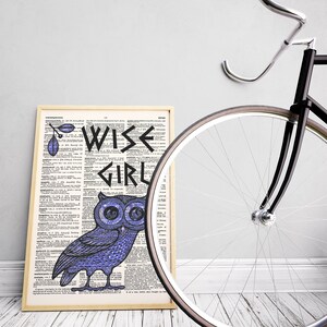May include: A framed print with a dictionary page background featuring a blue owl with the words "WISE GIRL" in black lettering. The print is leaning against a black bicycle with a white tire.