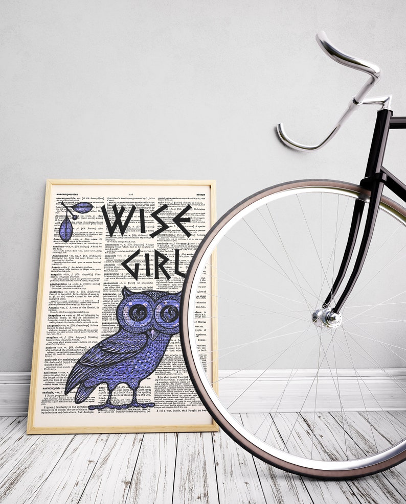May include: A framed print with a dictionary page background featuring a blue owl with the words "WISE GIRL" in black lettering. The print is leaning against a black bicycle with a white tire.