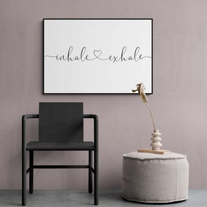 Pu&ograve; includere: Una stampa incorniciata nera con il testo "inhale &hearts; exhale" su sfondo bianco. La stampa &egrave; appesa a una parete rosa chiaro sopra una sedia nera e un pouf beige chiaro.