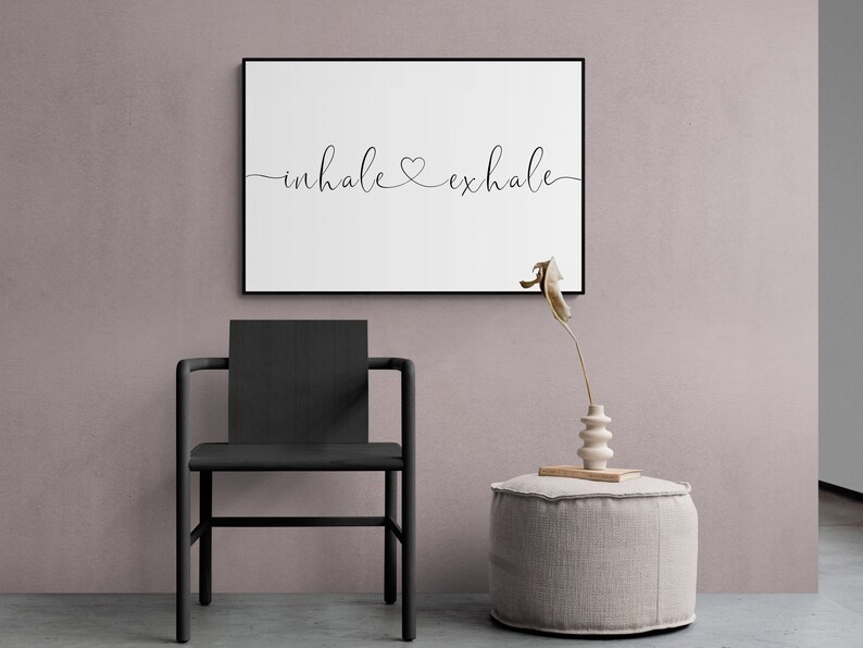 Pu&ograve; includere: Una stampa incorniciata nera con il testo "inhale &hearts; exhale" su sfondo bianco. La stampa &egrave; appesa a una parete rosa chiaro sopra una sedia nera e un pouf beige chiaro.