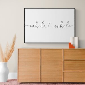 Pu&ograve; includere: Una stampa incorniciata con il testo "inhale &hearts; exhale" in scrittura corsiva nera su sfondo bianco. La stampa &egrave; appesa a una parete beige chiaro sopra un mobile in legno con tre cassetti.