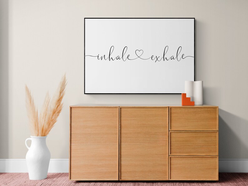 Pu&ograve; includere: Una stampa incorniciata con il testo "inhale &hearts; exhale" in scrittura corsiva nera su sfondo bianco. La stampa &egrave; appesa a una parete beige chiaro sopra un mobile in legno con tre cassetti.