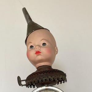 May include: A whimsical assemblage featuring a doll head with a metal funnel, a clock face, and a vintage tin. The clock face is set within a metal frame, and the base is a decorative black stand.