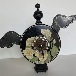 May include: A decorative clock with a black face featuring white floral designs and a crystal-studded center. The clock has metal wings and a black base and top, creating a unique, artistic piece.
