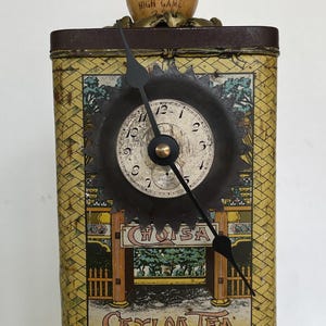 May include: A vintage clock made from a repurposed Ceylon Tea tin, featuring a bowling pin topper. The clock face is black with white numbers and hands. The base is a decorative brass trophy base. The clock is a unique decorative item.