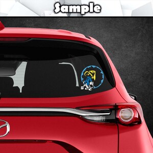 K&ouml;nnte beinhalten: Ein Autoaufkleber mit einem blau-gelben Cartoon-Charakter in einem blauen Kreis mit einem gelben Zahnraddesign. Der Aufkleber befindet sich auf der Heckscheibe eines roten Autos. Das Wort "Sample" ist oben aufgedruckt.