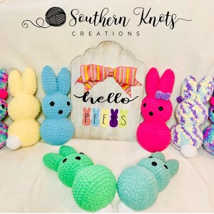 May include: A collection of colorful crocheted Easter bunny plushes. The bunnies are in various colors, including pink, blue, yellow, green, and purple. The bunnies are arranged in a line on a white surface. The sign behind the bunnies says "hello peeps".