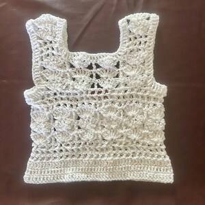 May include: A white crocheted sleeveless top with a delicate, lacy pattern. The top has a square neckline and a slightly cropped length.
