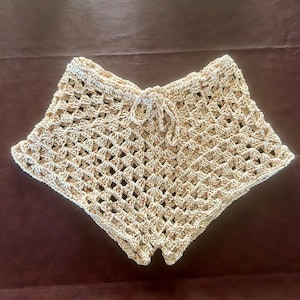 May include: A pair of beige crochet shorts featuring a diamond-shaped openwork pattern and a drawstring waistband. The yarn is a light tan color with subtle flecks of brown and orange throughout the woven fabric construction.