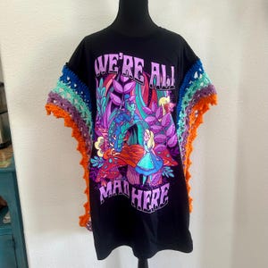 May include: A black t-shirt featuring a colorful graphic of Alice and the Caterpillar with the text "WE'RE ALL MAD HERE." The sleeves are accented with layers of vibrant, multi-colored crocheted trim in shades of blue, teal, purple, and orange.