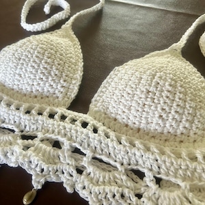 May include: A white crochet bikini top with a halter neck design. The top features a detailed crochet pattern and small wooden beads hanging from the bottom edge. The overall style is bohemian and handmade.