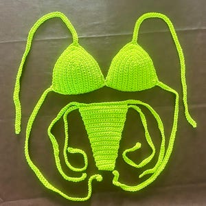May include: A neon green crochet bikini set consisting of a triangle top with thin tie straps and a matching bottom with side tie closures. The garment is crafted from a textured yarn material in a vibrant, solid lime green color throughout.