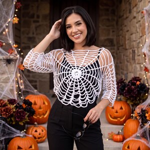 May include: A white crochet top featuring a spider web pattern with long sleeves and a round neckline, worn over a black camisole paired with black trousers. The garment has an open-knit design that radiates from the center of the chest.