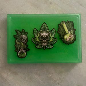 May include: A rectangular, translucent green tray with three stickers. The stickers feature skull designs, cannabis leaves, and a bong. The stickers have black outlines and are in shades of green, gray, and white.