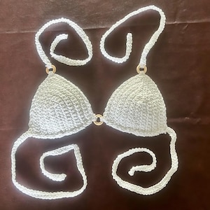 May include: A white crochet bikini top with triangle cups and adjustable straps. The top features gold-colored rings at the apexes and center. The crochet work has a textured appearance, and the straps are designed to tie around the neck and back.