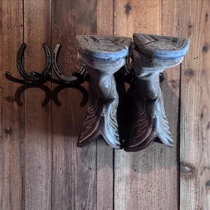 Puede incluir: Un par de botas de vaquero marrones expuestas en un estante para botas con forma de herradura. Las botas tienen costuras decorativas y están colocadas sobre un fondo de madera rústica. El estante es de metal negro.