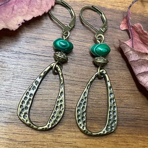 May include: A pair of handcrafted earrings with a teardrop shape. The earrings feature a hammered bronze finish, green malachite beads, and decorative metal accents. The lever-back closures are visible.