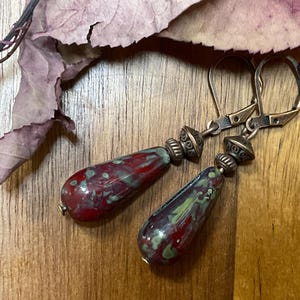 May include: A pair of dangle earrings featuring teardrop-shaped glass beads with a mottled pattern of deep red and sage green. Each bead is topped with a small, textured antique bronze-toned metal spacer and attached to a matching leverback hook.