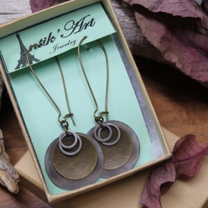 May include: A pair of dangle earrings featuring layered circular metal discs in bronze and dark gray tones, suspended from long curved ear wires. The earrings are presented in a light green cardboard box labeled Antik' Art Jewelry.