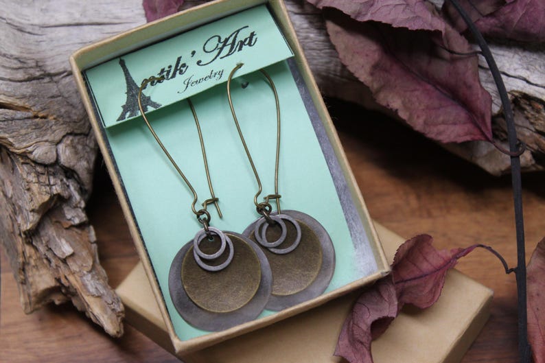 May include: A pair of dangle earrings featuring layered circular metal discs in bronze and dark gray tones, suspended from long curved ear wires. The earrings are presented in a light green cardboard box labeled Antik' Art Jewelry.