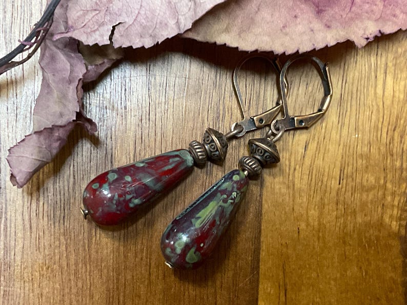 May include: A pair of dangle earrings featuring teardrop-shaped glass beads with a mottled pattern of deep red and sage green. Each bead is topped with a small, textured antique bronze-toned metal spacer and attached to a matching leverback hook.
