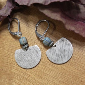 May include: Silver-tone earrings with a half-moon design and a textured finish. Each earring has a small, light blue and grey beaded detail and a lever-back clasp. The earrings are presented on a wooden surface with dried leaves.