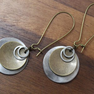 May include: A pair of dangle earrings featuring a large brushed silver-tone disc layered with a smaller antique brass-tone disc and two tiny silver-tone rings. The earrings are attached to long, thin, curved brass-tone ear wires for secure wear.