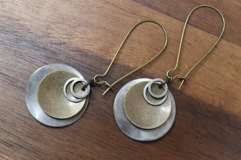 May include: A pair of dangle earrings featuring a large brushed silver-tone disc layered with a smaller antique brass-tone disc and two tiny silver-tone rings. The earrings are attached to long, thin, curved brass-tone ear wires for secure wear.