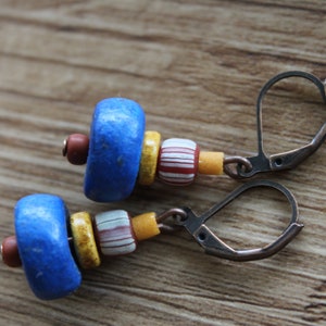 May include: A pair of handmade earrings featuring a combination of blue, yellow, red, and white beads. The earrings have a lever-back closure and are displayed on a wooden surface. The design includes a mix of shapes and colors.